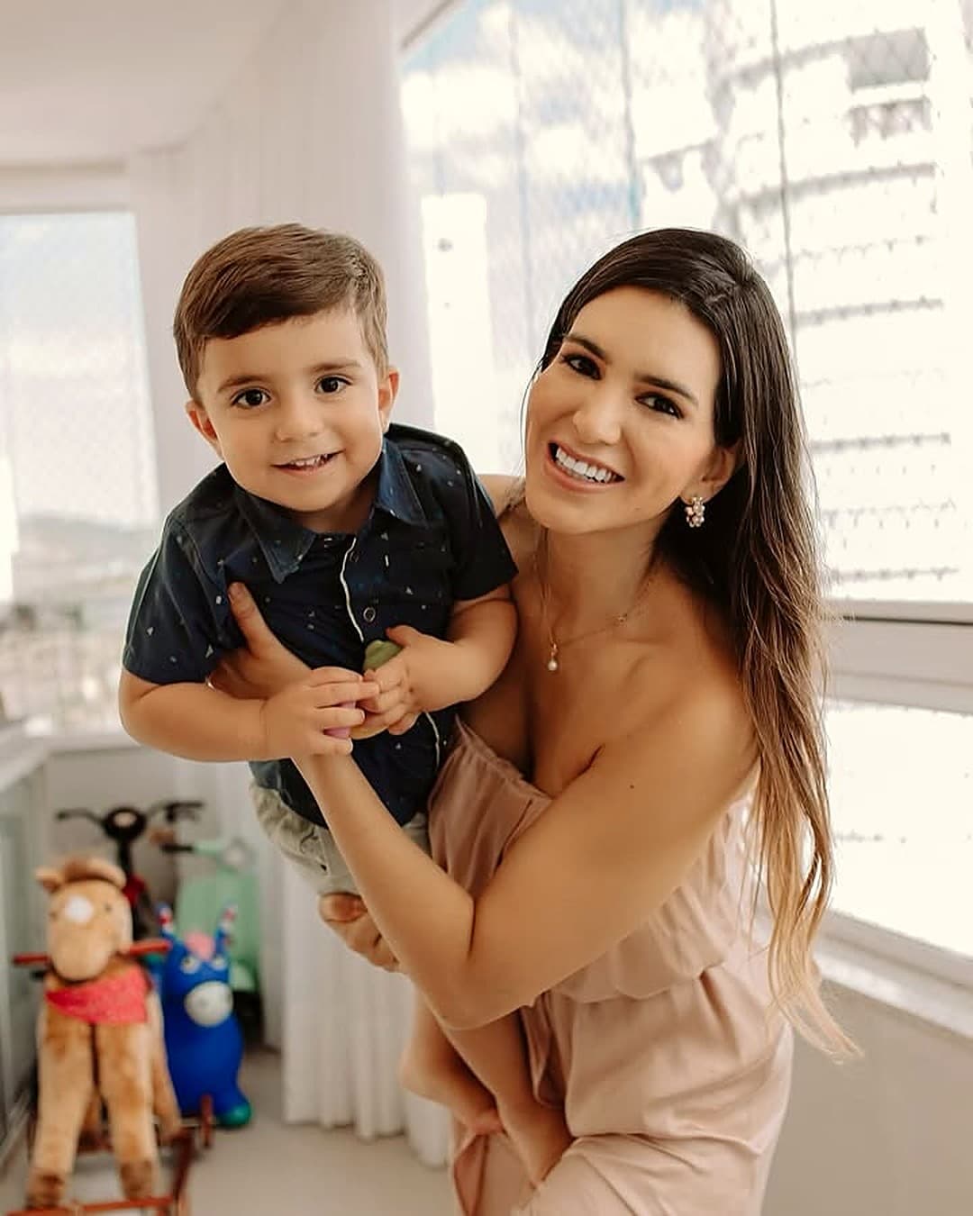 Dra. Raíssa Campelo sorrindo com uma criança no colo, em uma sala clara com brinquedos ao fundo